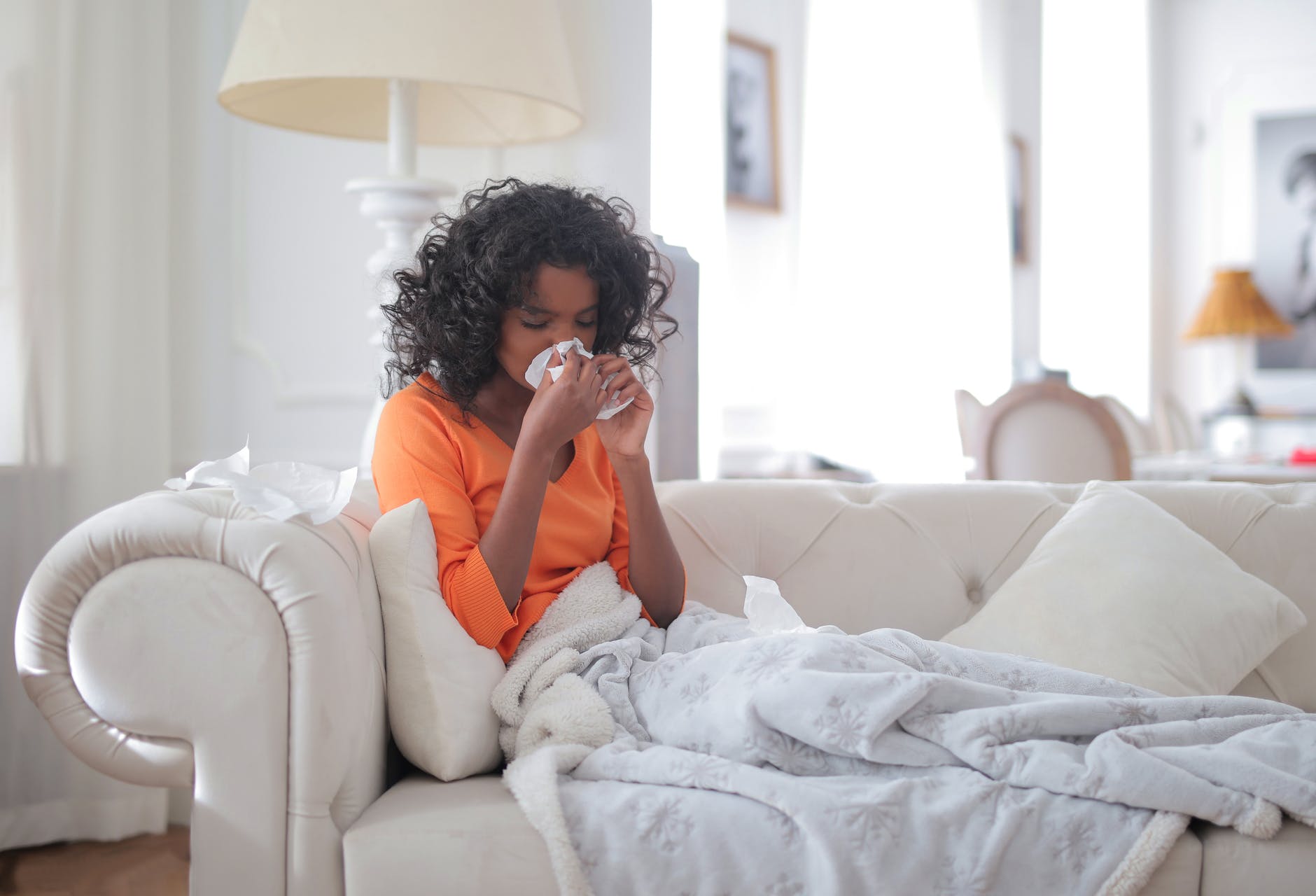Woman in orange top sitting on with couch, covered by orane blanket, wiping her nose with a tissue.